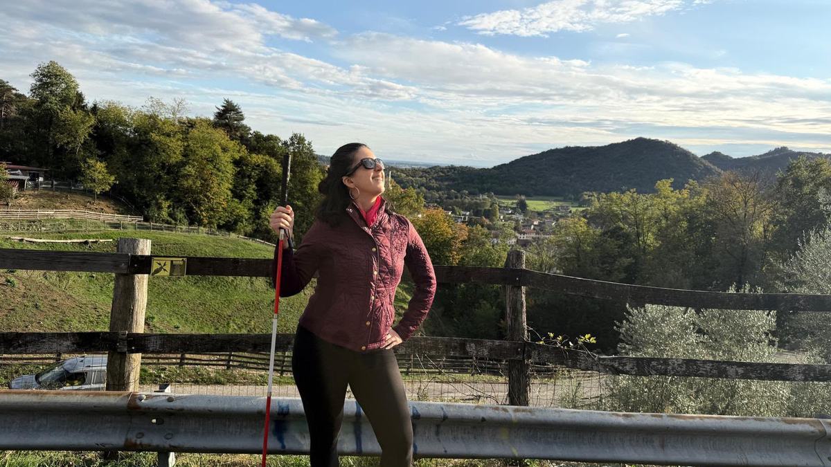 Catarina Rivera, una giovane donna con i capelli castani, un bastone bianco e rosso e gli occhiali da sole, sorride con le montagne sullo sfondo.